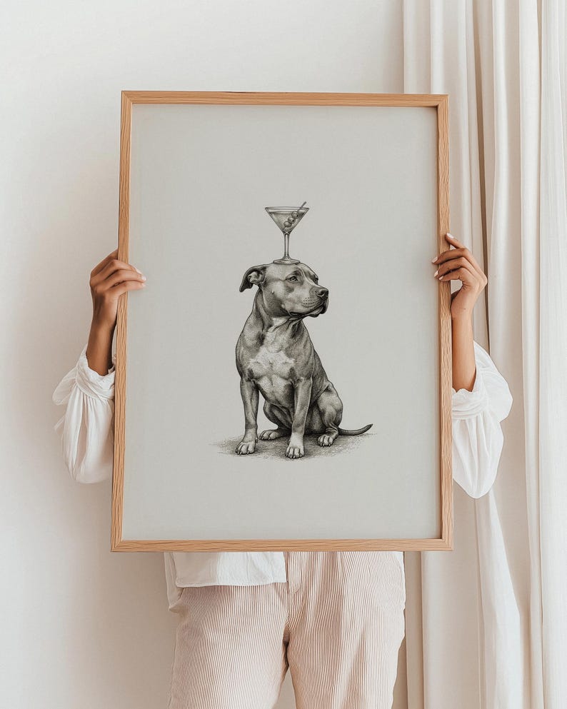 May include: A framed print featuring a detailed pencil drawing of a seated pit bull with a martini glass balanced on its head. The artwork is in grayscale, with a light wooden frame. The background is a neutral, off-white color.