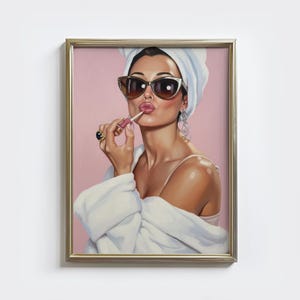 Może przedstawiać: Oprawione dzieło sztuki przedstawiające kobietę nakładającą szminkę, noszącą okulary przeciwsłoneczne, biały ręcznik i biały szlafrok. Tło jest w kolorze miękkiego różu. Kobieta ma na sobie duże kolczyki koła i pierścionek. Rama jest złota.