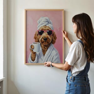 以下が含まれることがあります： サングラス、タオル、バスローブ姿の犬がマグカップを持っているフレーム入りプリント。犬はピンク色の背景に描かれています。フレームは明るい茶色です。