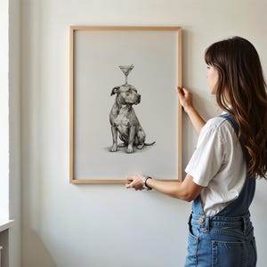 May include: A framed print featuring a detailed black and white illustration of a dog with a martini glass balanced on its head. The artwork is in a light wooden frame, ready to hang. The print is displayed on a white wall.