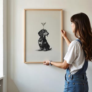 May include: A framed art print showing a monochrome illustration of a dachshund with a martini glass balanced on its head. The print is in a light wood frame and is being held against a white wall.