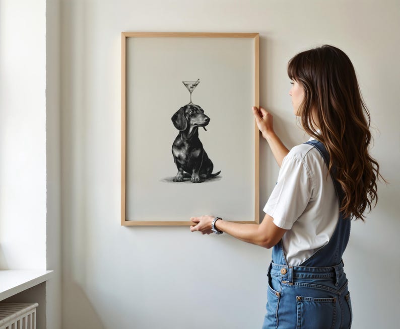 May include: A framed art print showing a monochrome illustration of a dachshund with a martini glass balanced on its head. The print is in a light wood frame and is being held against a white wall.