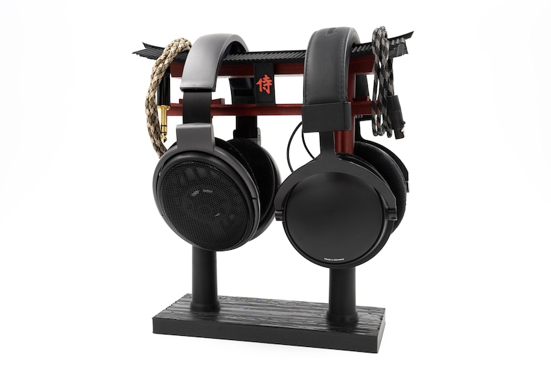 May include: Two black headphones with black stands on a wooden base. The headphones are resting on a red and black stand with the Japanese character "信" written on it.