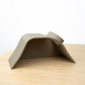 Pu&ograve; includere: Un supporto per telefono in silicone grigio, curvo, con un bordo rialzato sul retro. Il supporto &egrave; progettato per tenere un telefono in posizione verticale.
