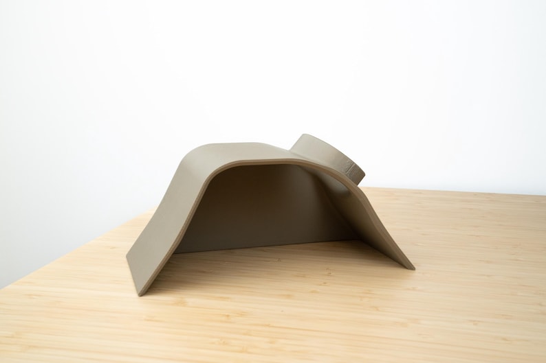 Pu&ograve; includere: Un supporto per telefono in silicone grigio, curvo, con un bordo rialzato sul retro. Il supporto &egrave; progettato per tenere un telefono in posizione verticale.