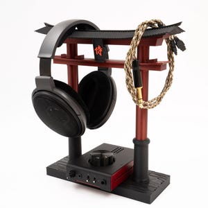 May include: A black headphone stand with a red wooden torii gate design. The stand has a black base and a black headphone holder. A black and gold headphone cable is wrapped around the torii gate.