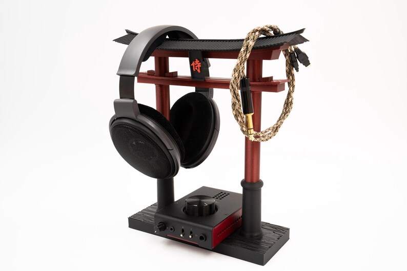 May include: A black headphone stand with a red wooden torii gate design. The stand has a black base and a black headphone holder. A black and gold headphone cable is wrapped around the torii gate.