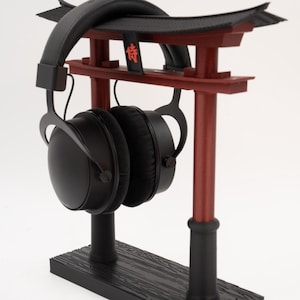 May include: Black headphones with a black and red torii gate stand. The stand has a wooden base and the torii gate has the kanji character for "lucky" written on it.