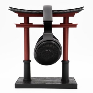 May include: Black headphones hanging from a red and black wooden torii gate stand. The stand has a black base with a wood grain pattern.