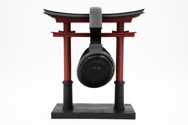 May include: Black headphones hanging from a red and black wooden torii gate stand. The stand has a black base with a wood grain pattern.