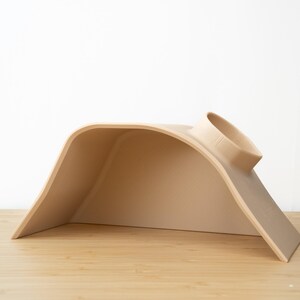 Pu&ograve; includere: Un letto per gatti in plastica beige a forma di tunnel curvo con un'apertura rotonda nella parte superiore.