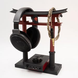 May include: A black headphone stand with a red wooden torii gate design. The stand features a black headphone holder and a red amplifier with a knob. The stand also includes a black and beige braided cable.