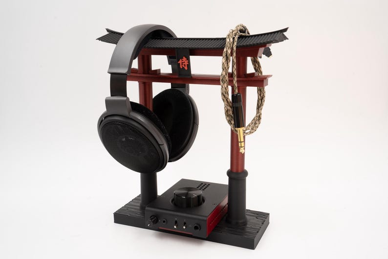May include: A black headphone stand with a red wooden torii gate design. The stand features a black headphone holder and a red amplifier with a knob. The stand also includes a black and beige braided cable.