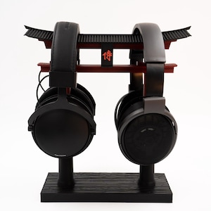 May include: Two pairs of black headphones are displayed on a black stand with a red and black Japanese-style gate design. The gate has the Japanese character "侍" (Samurai) written on it.