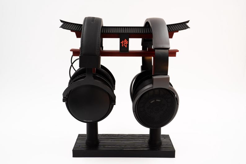 May include: Two pairs of black headphones are displayed on a black stand with a red and black Japanese-style gate design. The gate has the Japanese character "侍" (Samurai) written on it.