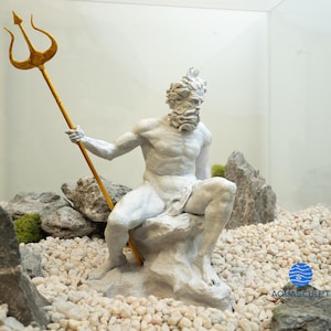 以下が含まれることがあります： 金色の三叉槍を持った神話上のキャラクターの白い塗装された像。像は白い小石で囲まれた岩の上に座っています。背景は白い壁とガラスケースです。