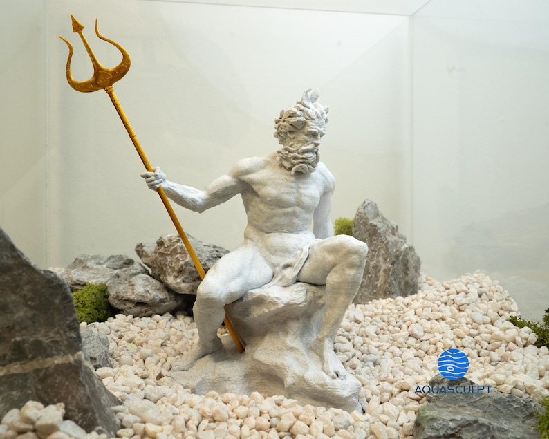 以下が含まれることがあります： 金色の三叉槍を持った神話上のキャラクターの白い塗装された像。像は白い小石で囲まれた岩の上に座っています。背景は白い壁とガラスケースです。