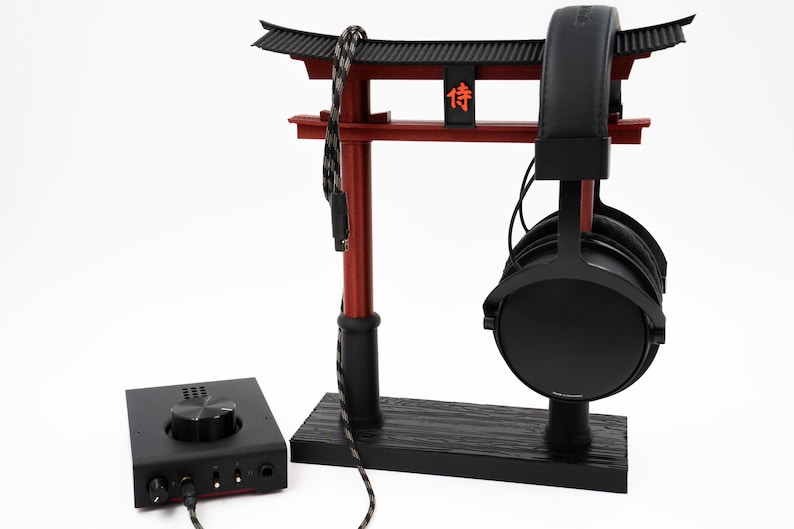 May include: Black headphones with a black leather headband are displayed on a black and red wooden stand shaped like a Japanese torii gate. The stand has the Japanese character "侍" written on it. A black and red audio amplifier is in the foreground.