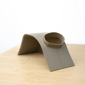 Pu&ograve; includere: Un supporto per telefono in silicone beige con un design curvo e un bordo rialzato per tenere un telefono in posizione.