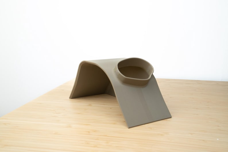 Pu&ograve; includere: Un supporto per telefono in silicone beige con un design curvo e un bordo rialzato per tenere un telefono in posizione.