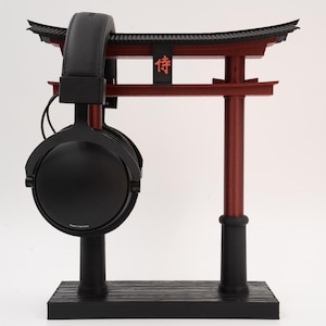 May include: Black headphones with a black stand and a red torii gate. The torii gate has a black roof and the word "侍" in red.