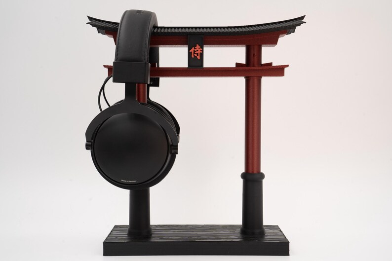May include: Black headphones with a black stand and a red torii gate. The torii gate has a black roof and the word "侍" in red.