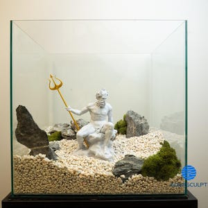 以下が含まれることがあります： 金色の三叉槍を持った神話上の海の神の白い像が、白い砂利、苔、灰色の岩で満たされたガラスのテラリウムの中に置かれています。テラリウムは黒い台座の上にあります。
