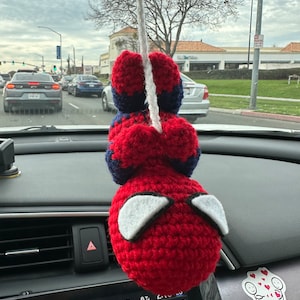 May include: A red and blue crocheted Spiderman hanging from a rearview mirror in a car. The car's dashboard is visible in the foreground, with a sticker of two stick figures holding hearts.