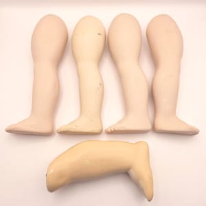 May include: Five antique doll limbs are arranged against a white backdrop. The collection includes four legs and one arm, all crafted from a pale, flesh-toned material. The legs show detailed feet and knees, while the arm is bent at the elbow.