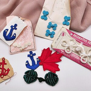 May include: A selection of vintage sewing accessories, featuring blue anchor appliques, red and green decorative frog closures, and blue bow-shaped buttons. The items are arranged on a white surface with a pink fabric backdrop.