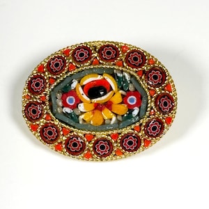 May include: An oval mosaic brooch with a gold-tone frame. The mosaic features a central flower design with yellow petals and a black centre, surrounded by red and orange floral patterns. The brooch is set against a white background.
