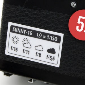 Puede incluir: Una cámara negra con una pegatina blanca que dice "SUNNY-16 ≈ 1/ISO" y muestra diferentes ajustes de apertura para diferentes condiciones de iluminación. La pegatina también incluye iconos para un día soleado, un día parcialmente nublado, un día nublado y un día nublado. Un círculo rojo con el texto "5x" es visible en la cámara.