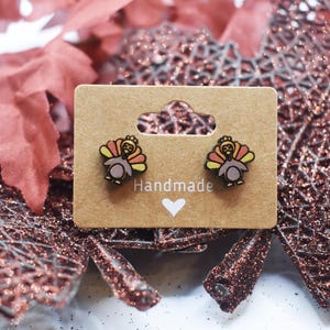 May include: A pair of handmade turkey earrings on a card with the word "Handmade" printed on it. The earrings are made of wood and painted in shades of orange, yellow, and brown.