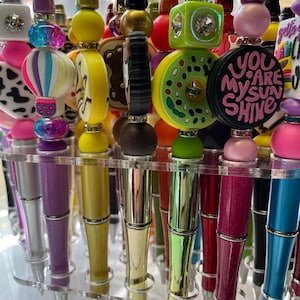 May include: A display of colorful pens with various decorative toppers, including a donut, a hot air balloon, a cow print, and a black and pink charm with the text "You Are My Sunshine".