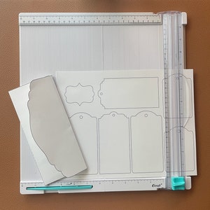 Op de afbeelding: Een witte snijmat met een liniaal en een set papieren sjablonen voor het maken van labels en enveloppen. De sjablonen omvatten een verscheidenheid aan vormen en maten, waaronder rechthoekige, ovale en hartvormige labels. De mat is gemarkeerd met inches en heeft een rasterpatroon.