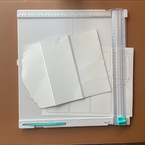 Op de afbeelding: Een witte snijmat met een liniaal en een snijgereedschap. De mat heeft een rasterpatroon en wordt gebruikt voor knutselen en scrapbooking. Er ligt een witte papieren sjabloon op de mat.