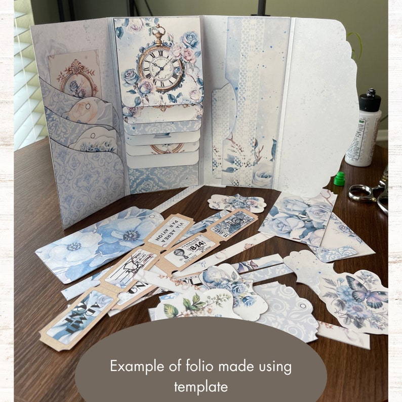 K&ouml;nnte beinhalten: Ein Beispiel f&uuml;r ein Folio, das mit einer Vorlage erstellt wurde. Das Folio besteht aus wei&szlig;em Karton mit blauen und wei&szlig;en Blumenmustern. Das Folio ist ge&ouml;ffnet und das Innere ist mit verschiedenen Etiketten, Tickets und anderen Verzierungen dekoriert. Das Folio ist eine gro&szlig;artige M&ouml;glichkeit, Fotos, Erinnerungsst&uuml;cke und andere Andenken aufzubewahren und auszustellen.