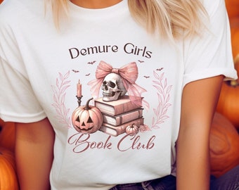 May include: A white t-shirt with a pink and black skull and books graphic. The text "Demure Girls Book Club" is printed on the shirt.