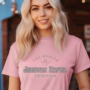 May include: A pink t-shirt with the text "The Healer Jehovah Rapha Exodus 15:26" in a gray font. The text is in a circle with a stylized image of a person with arms raised.