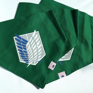 Könnte beinhalten: Drei grüne Stoffbandanas mit einem weißen und blauen geflügelten Emblem, das darauf gedruckt ist. Die Bandanas sind gefaltet und übereinander gestapelt. Die Bandanas haben ein rosa Etikett mit dem Wort "MANKAI" darauf gedruckt.