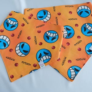 Op de afbeelding: Drie oranje bandana-slabbetjes met een blauw lachend gezichtpatroon. De bandana's hebben de tekst "3387" en "AXCE" erop gedrukt.