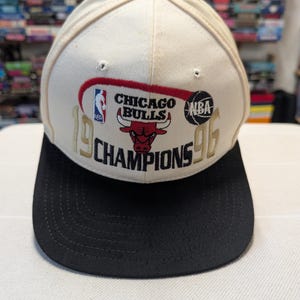 May include: A cream-colored Chicago Bulls baseball cap with a black brim. The cap features the team logo, the NBA logo, and the words "CHICAGO BULLS CHAMPIONS 96" in gold and red.