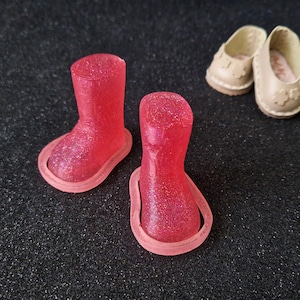 May include: Two pairs of miniature doll shoes. One pair of red glitter boots and one pair of beige flats with flower details. The boots are knee-high with a rounded toe. The flats have a rounded toe and a small heel.