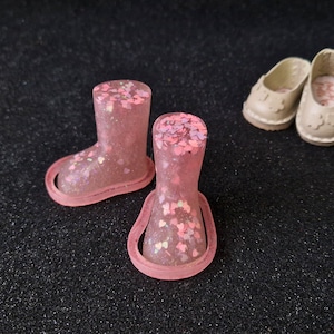 May include: Two pink glittery doll boots and a pair of beige doll shoes are displayed on a black surface. The boots have a rounded toe and a strap around the ankle. The shoes have a rounded toe and a scalloped edge.