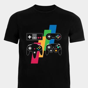 May include: Black t-shirt featuring a colourful graphic of four different video game controllers. The controllers are from different generations of gaming consoles, including the Nintendo Entertainment System, Super Nintendo Entertainment System, Nintendo 64, and Sega Genesis.