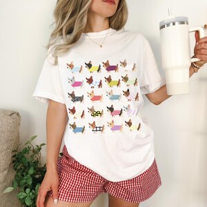 May include: Ivory-colored t-shirt featuring a repeating pattern of dachshund dogs wearing various outfits. The dogs are in different colors and patterns. The model is wearing red and white checkered shorts and holding a white tumbler.