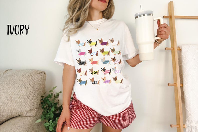 May include: Ivory-colored t-shirt featuring a repeating pattern of dachshund dogs wearing various outfits. The dogs are in different colors and patterns. The model is wearing red and white checkered shorts and holding a white tumbler.