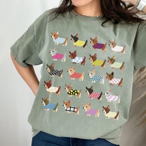 May include: Moss green t-shirt featuring a repeating pattern of cartoon dachshunds wearing various outfits. The word "MOSS" is printed in black script in the upper left corner. The shirt is a relaxed fit.