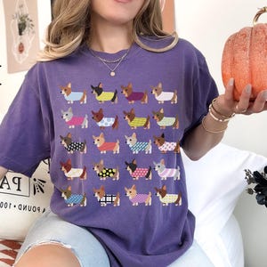 May include: A purple t-shirt with a repeating pattern of cartoon dachshunds wearing various outfits. The word "GRAPE" is printed in large letters on the upper left. A person is holding an orange pumpkin.