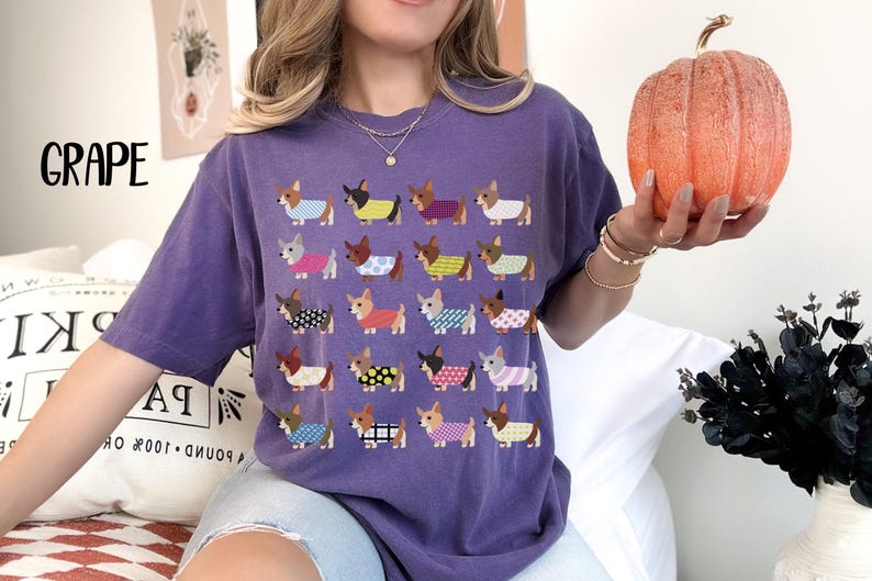 May include: A purple t-shirt with a repeating pattern of cartoon dachshunds wearing various outfits. The word "GRAPE" is printed in large letters on the upper left. A person is holding an orange pumpkin.